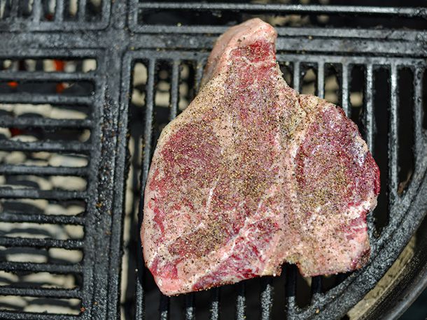 A raw, seasoned porterhouse has been placed on the cooler side of the grill and oriented so that the tenderloin is further away from the coals than the strip loin.