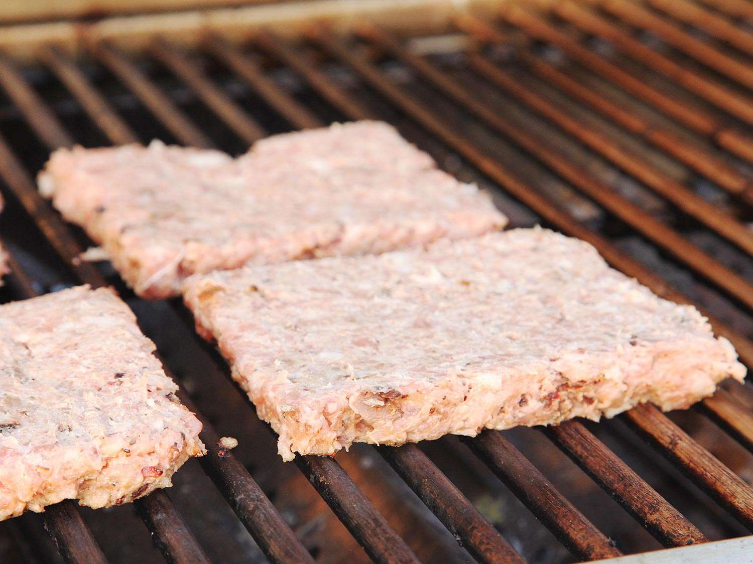 Three uncooked square rib patties on a grill.