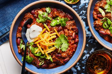 2 bowls of chili in blue bowls, on a blue marble surface, on a white tabletop with small dish of limes, green onions and a drink