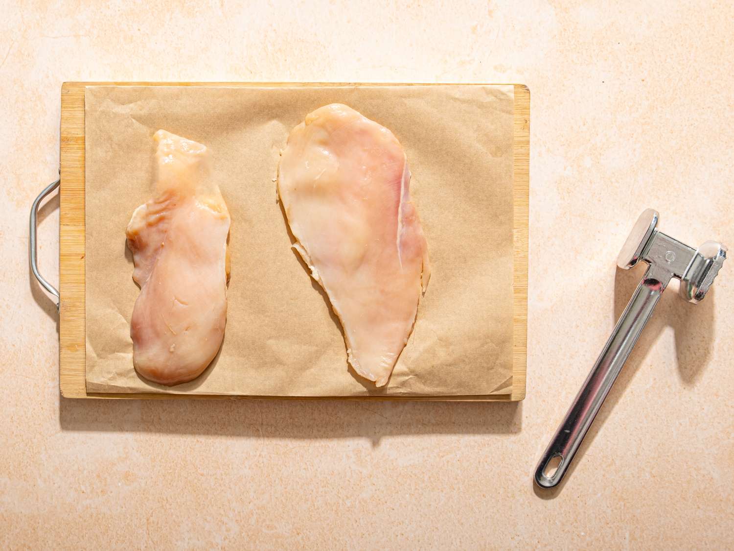 Two raw chicken breasts on a wooden cutting board a meat tenderizer placed on the side