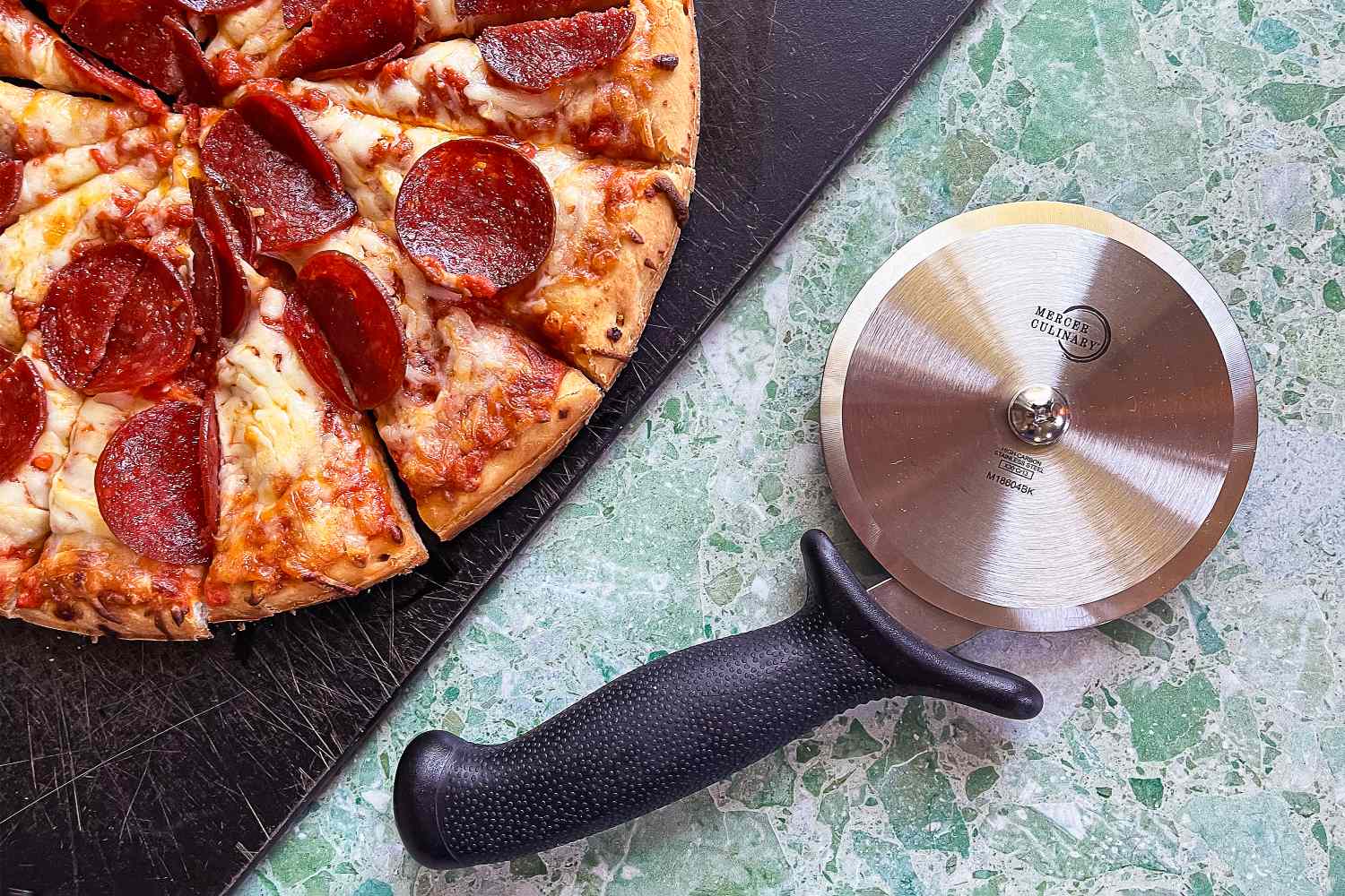 A pepperoni pizza on a black tray beside a Mercer stainless steel pizza cutter on a green countertop