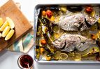 Overhead shot of 2 whole porgy roasted on a sheet pan with tomatoes, lemon slices, and shishito peppers