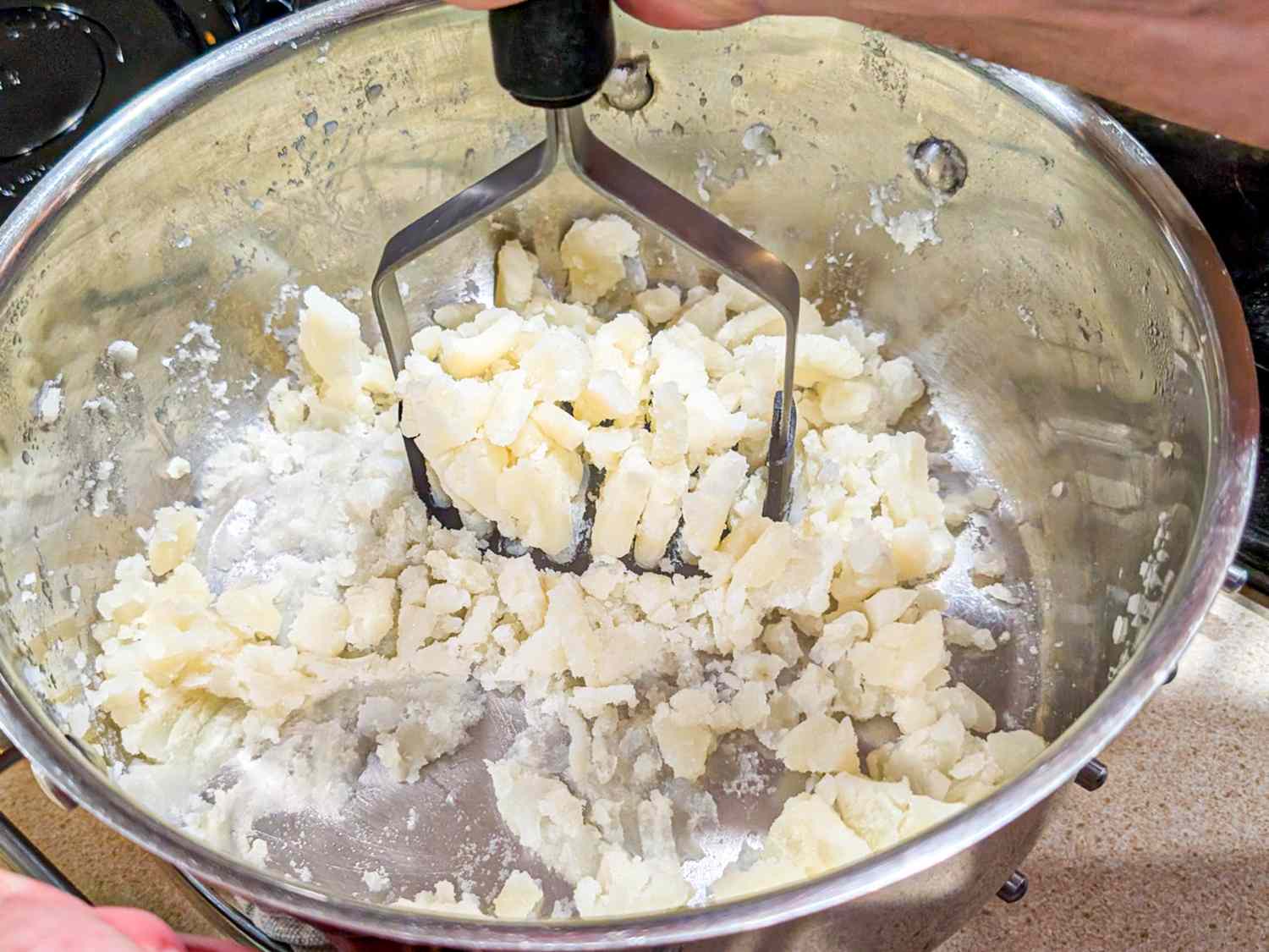 A metal potato masher pressing into a pot of partially mashed potatoes