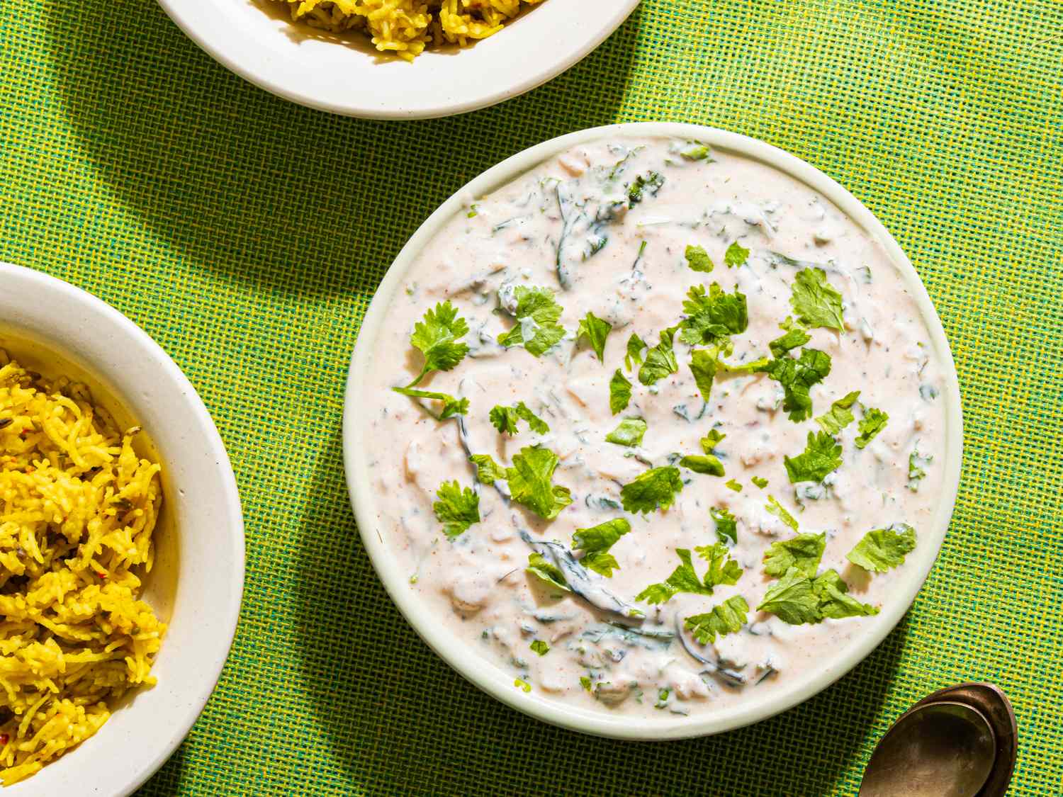 A bowl of raita garnished with herbs next to two bowls of spiced rice and a spoon