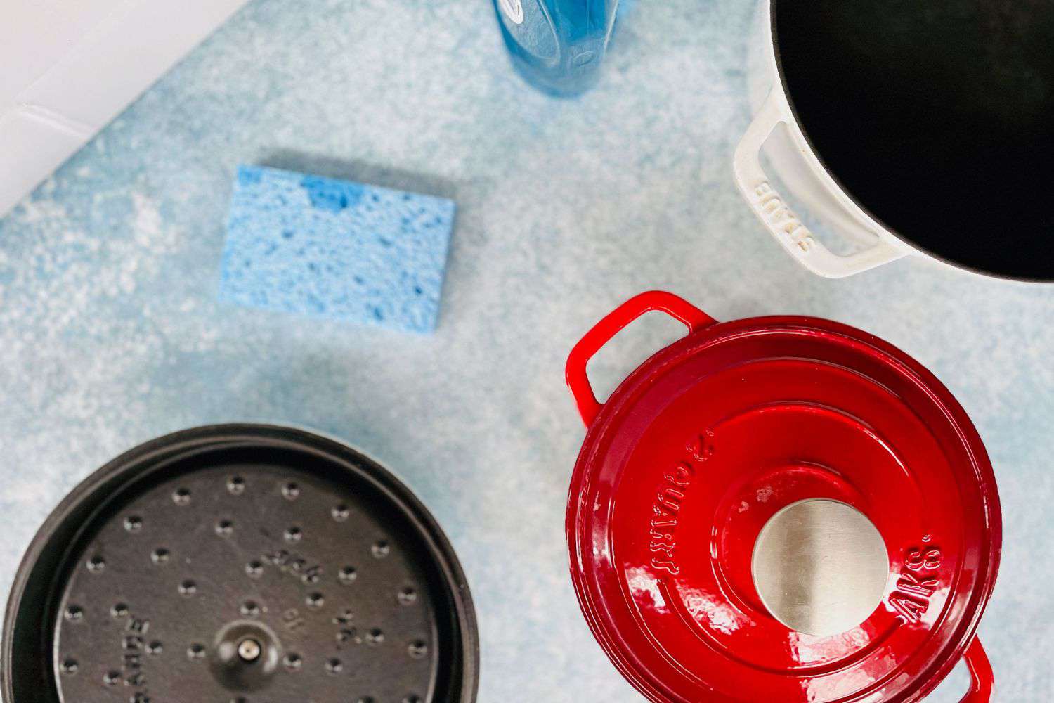 Three Dutch ovens sit on a countertop next to a sponge and dish soap.
