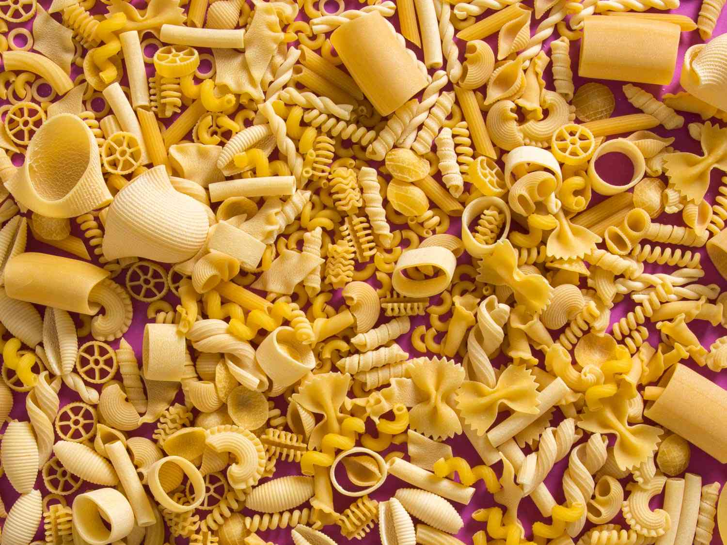 Dried pasta in various shapes against a pink background.