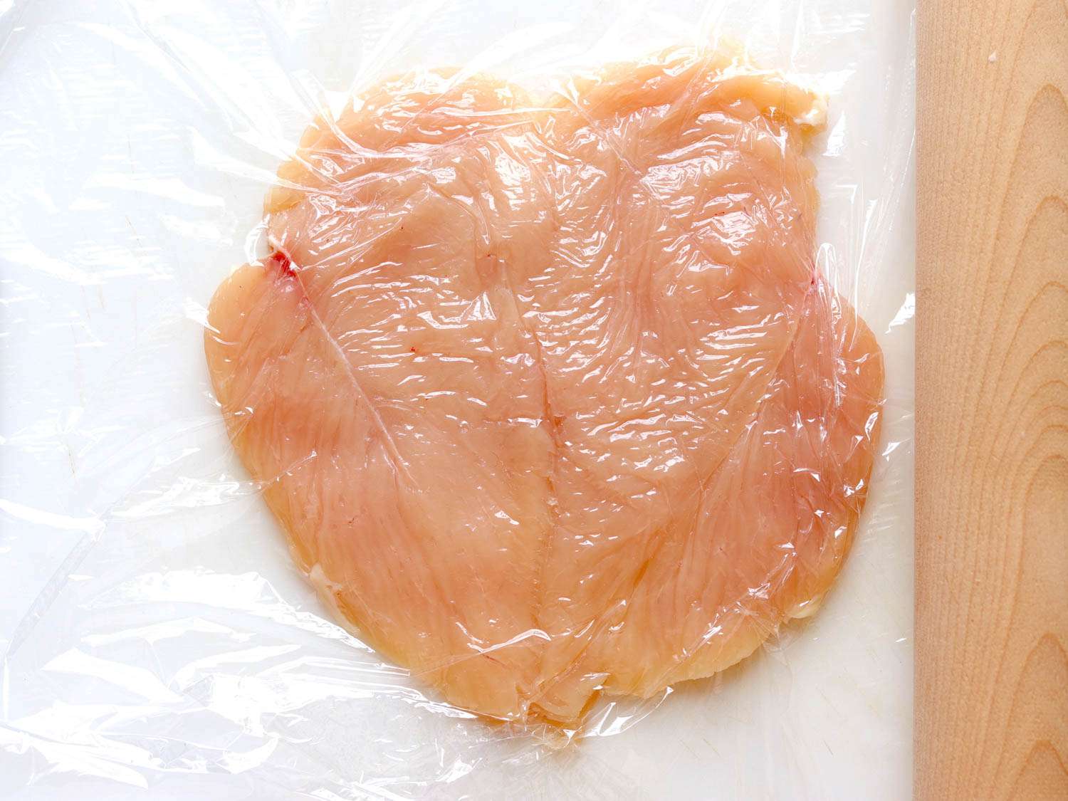 Skinless chicken breasts between plastic wrap, pounded thin with a wooden rolling pin.