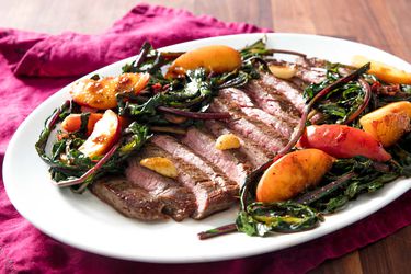 Closeup of Pan-Seared Flank Steak With Peaches and Dandelion Greens, served on a white oval platter.
