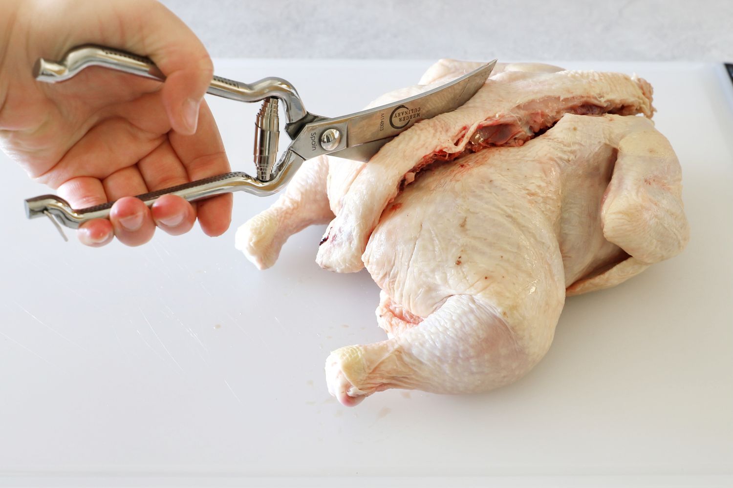 A hand holding Mercer Culinary poultry shears trimming a raw chicken