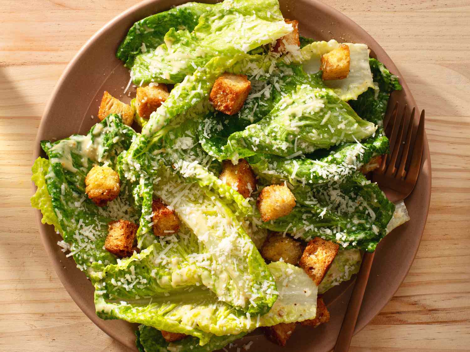 Composed caesar salad on a ceramic plate with a copper-colored fork, placed on a wooden table.