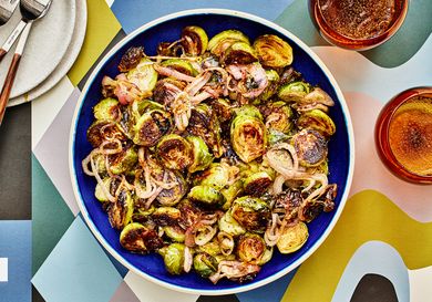 Roasted brussel sprouts and shallots with a balsamic glaze in a bowl, with glasses of sparkling water, some smaller plates and a colorfully printed table top