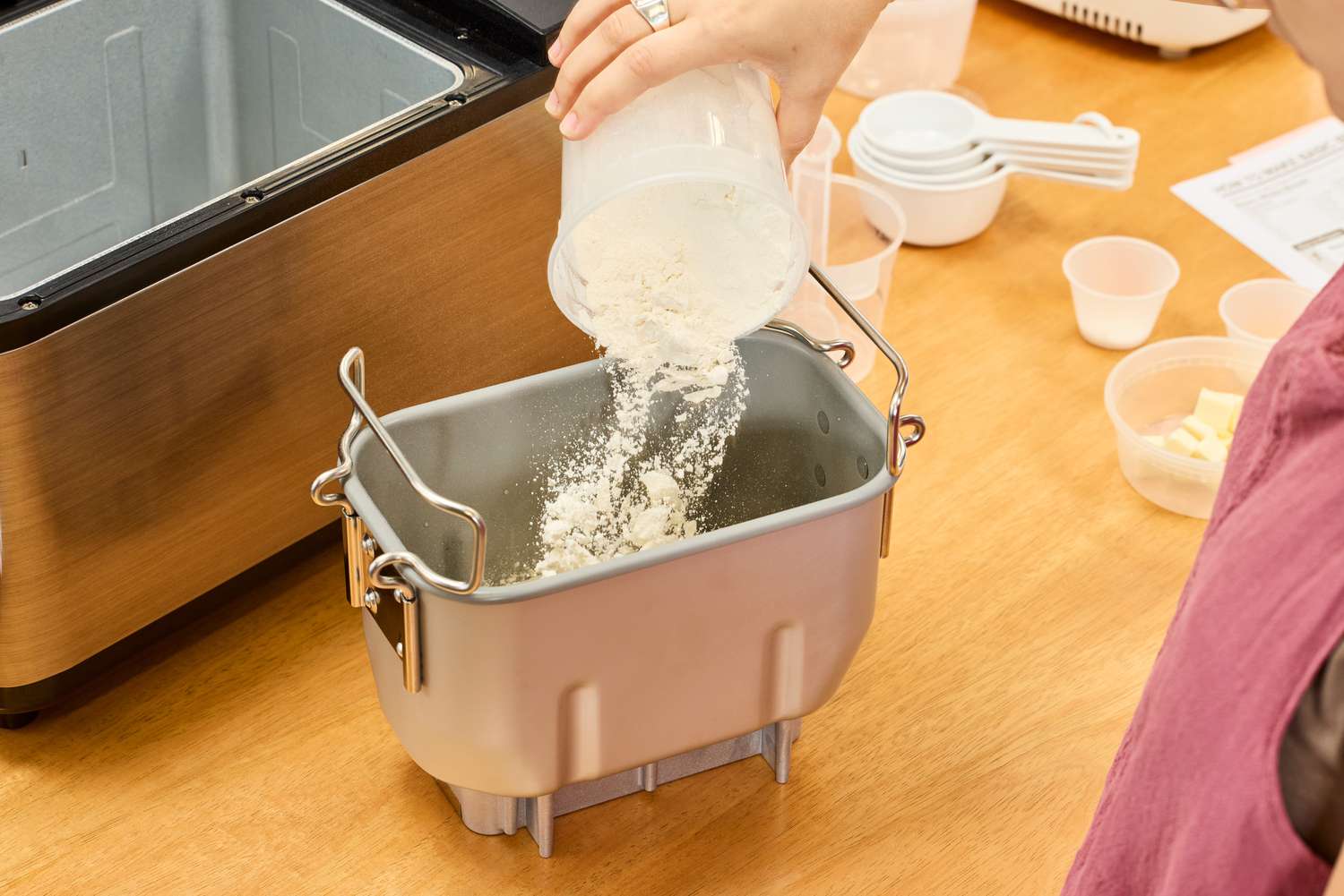 A hand adding flour into the Zojirushi Home Bakery Virtuoso Plus Bread Maker basket on wood