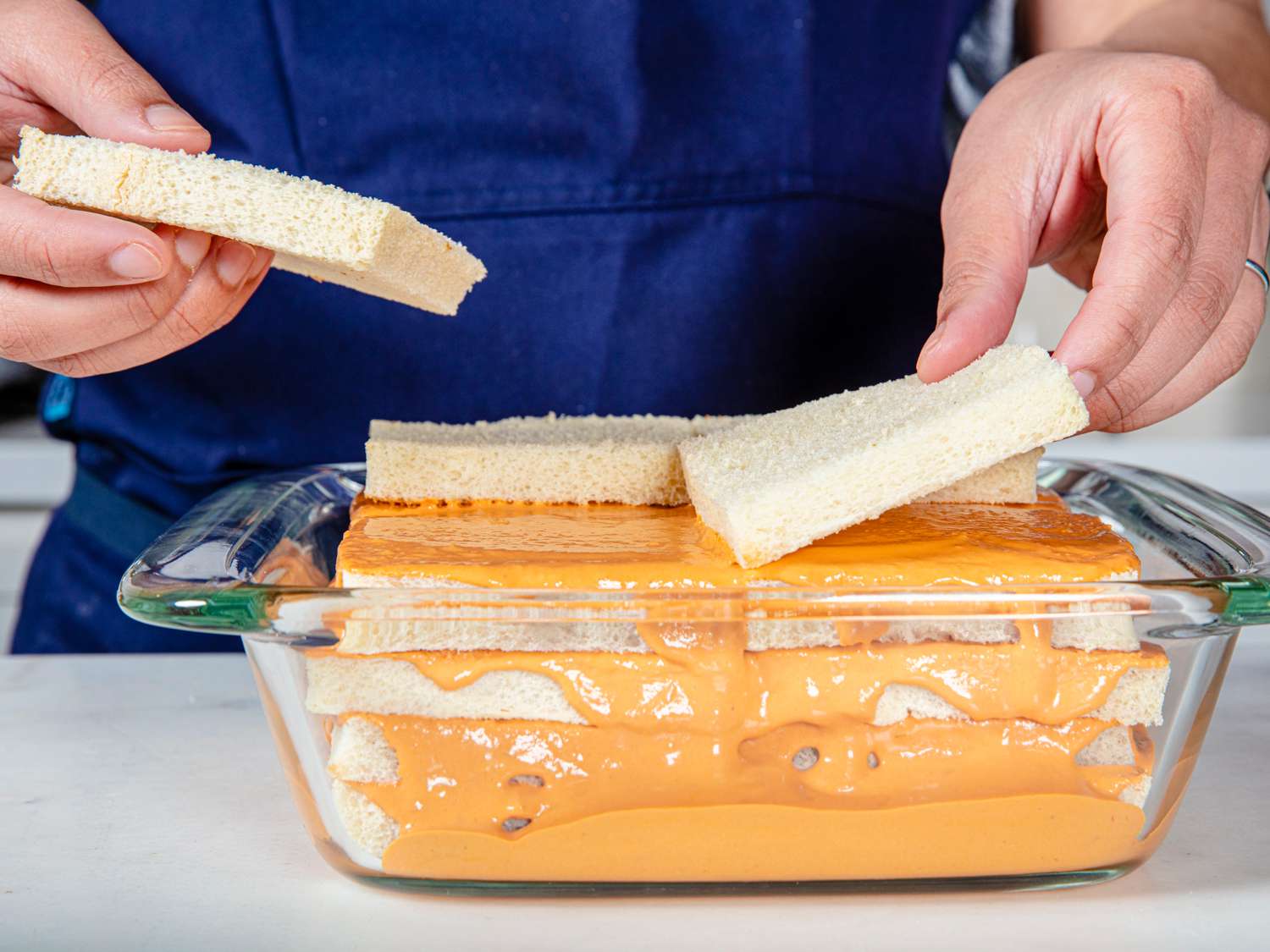 Angled view of a chef in a blue apron placing a layer of white bread into the pastel de atun