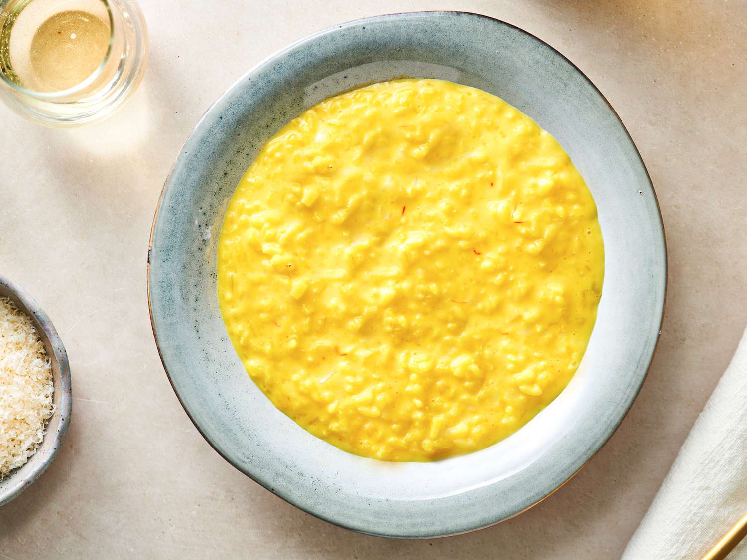 A round ceramic plate holding creamy saffron risotto. There is a glass of liquid in the top left corner and a small bowl of grated cheese on the bottom left edge of the frame.