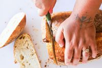 A person slicing bread with a Material knife on a cutting board hand with a ring visible