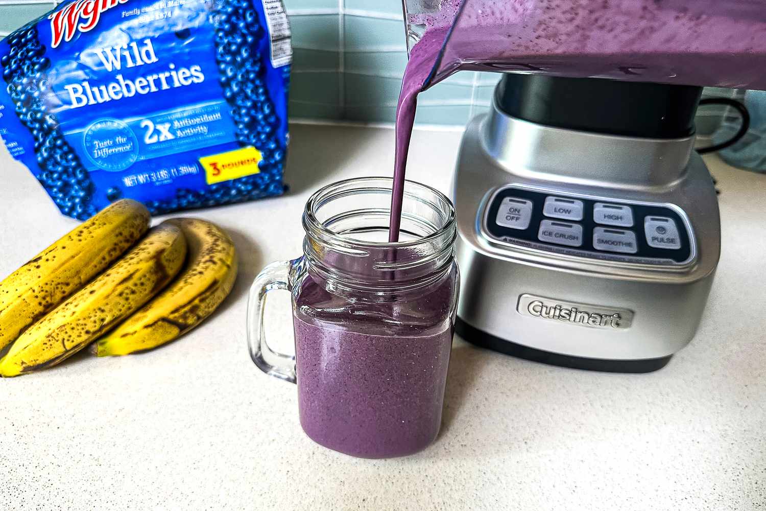 A blueberry and banana smoothie being poured from a blender jar into a glass on a kitchen counter