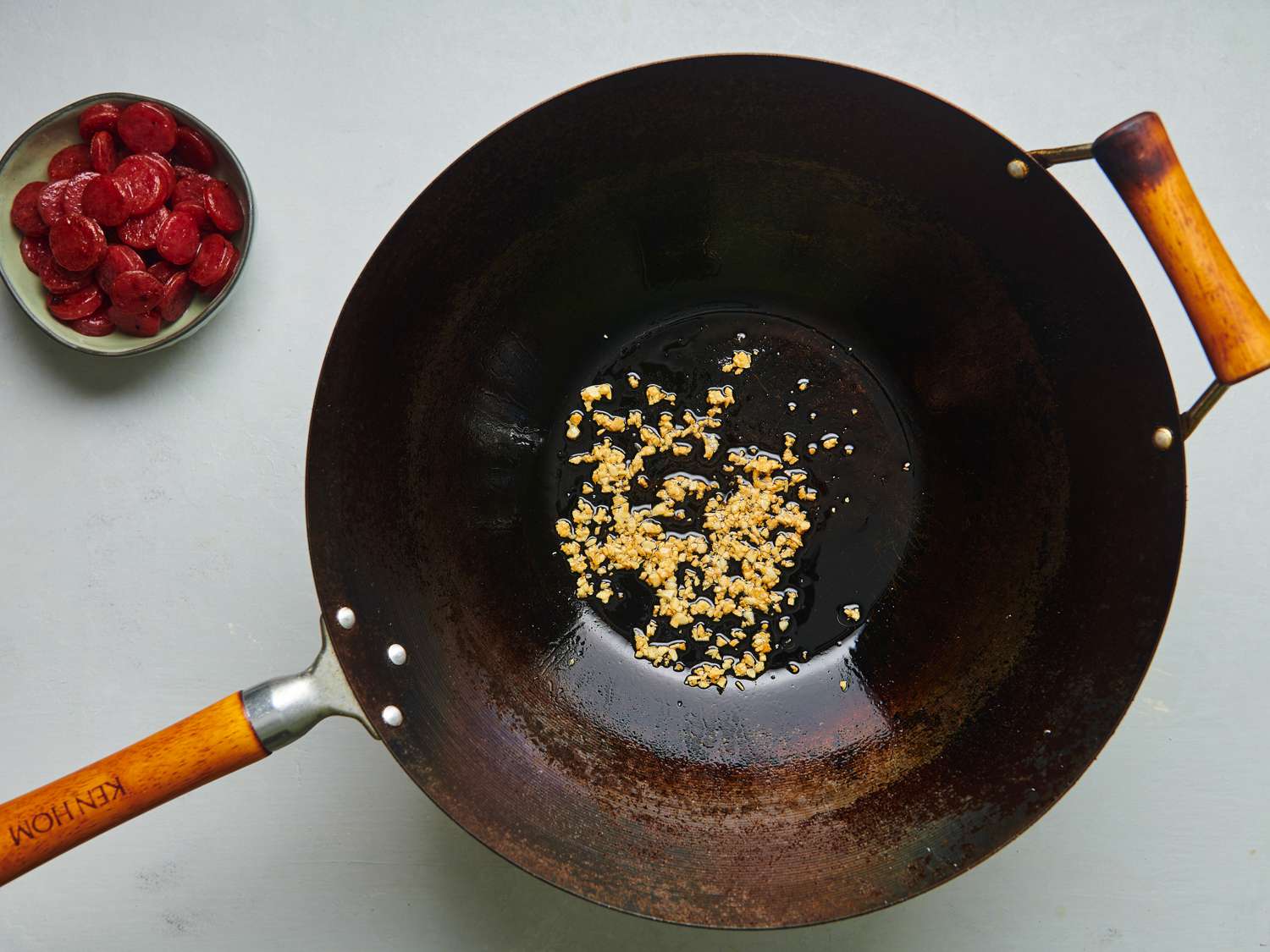 A well-seasoned carbon steel wok holding golden browned garlic and rendered fat from the Chinese sausage. The fried Chinese sausage is in a small bowl in the top left corner of the image.