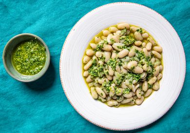 Overhead view of garlic scape pesto in a small bowl next to a wide rimmed, shallow bowl of cooked white beans and pesto