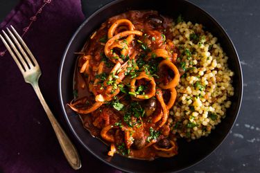 Overhead view of a bowl of braised squid with tomato, harissa, and olives, served with fregola pasta.
