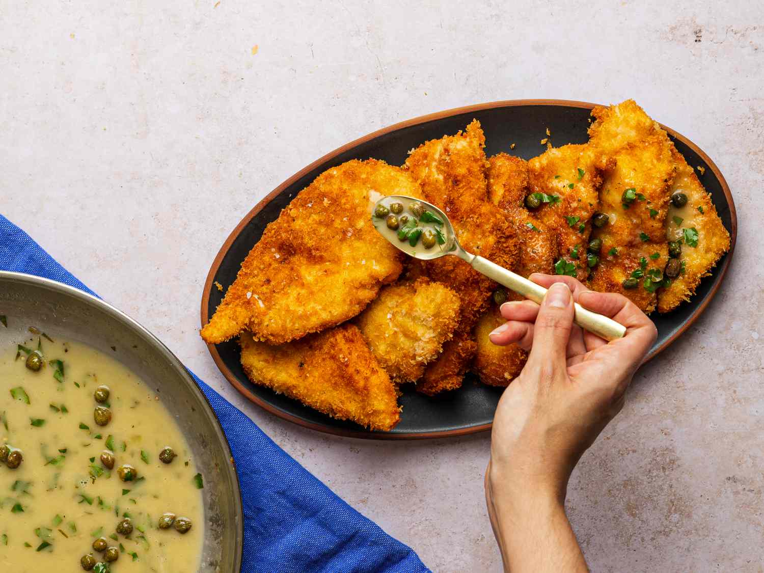 An oval platter covered with fried chicken cutlets. There is a hand holding a spoonful of the lemon-butter sauce, which is being distributed over the surface of the chicken.