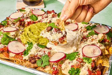 Nachos in foil, with a hand dipping a chip into toppings on a green surface, with a drink in the background