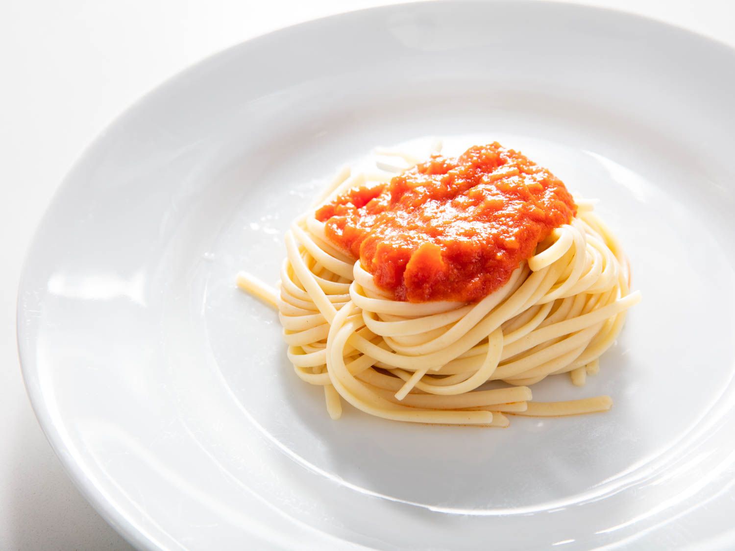 A pile of plain spaghetti sits on a plate with a splotch of sauce spooned on top