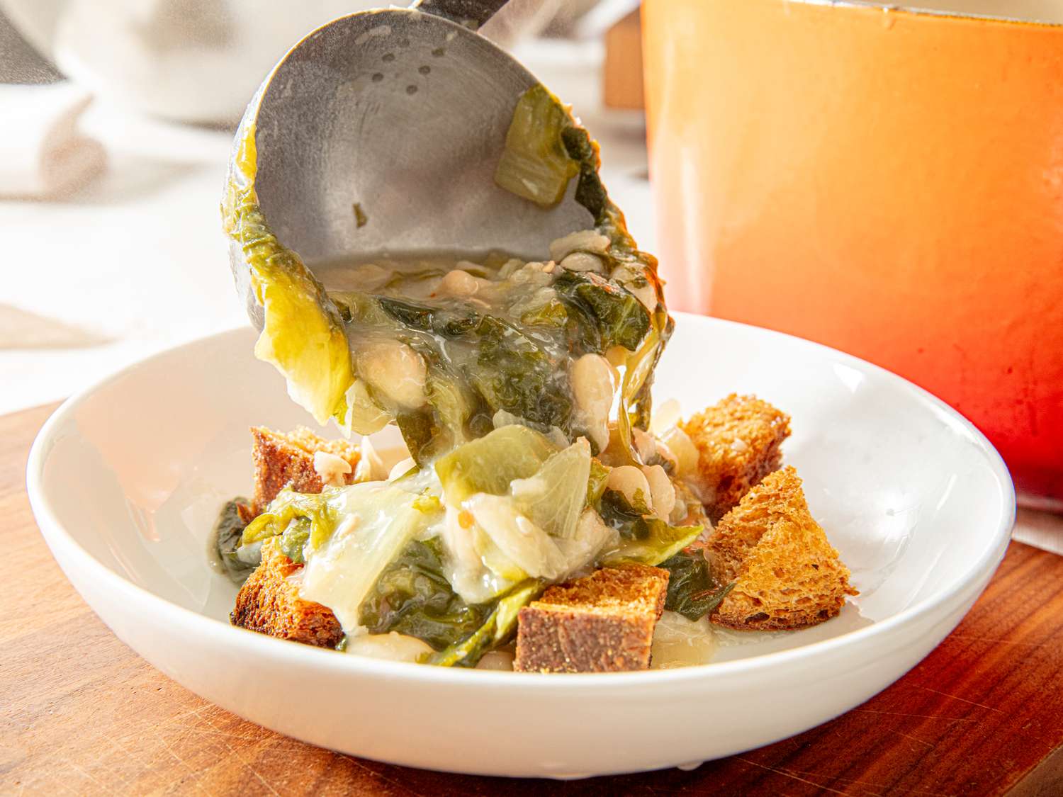 A bowl with croutons and a ladle pouring a soup containing leafy greens and beans