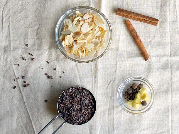 An assemblage of ingredients, including sliced almonds, cinnamon sticks, spices, and a measuring cup of cocoa nibs.