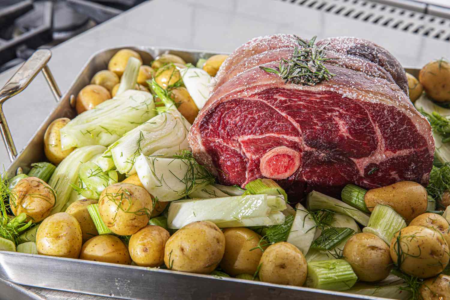 An uncooked roast surrounded by potatoes and fennel and situated in a roasting pan.