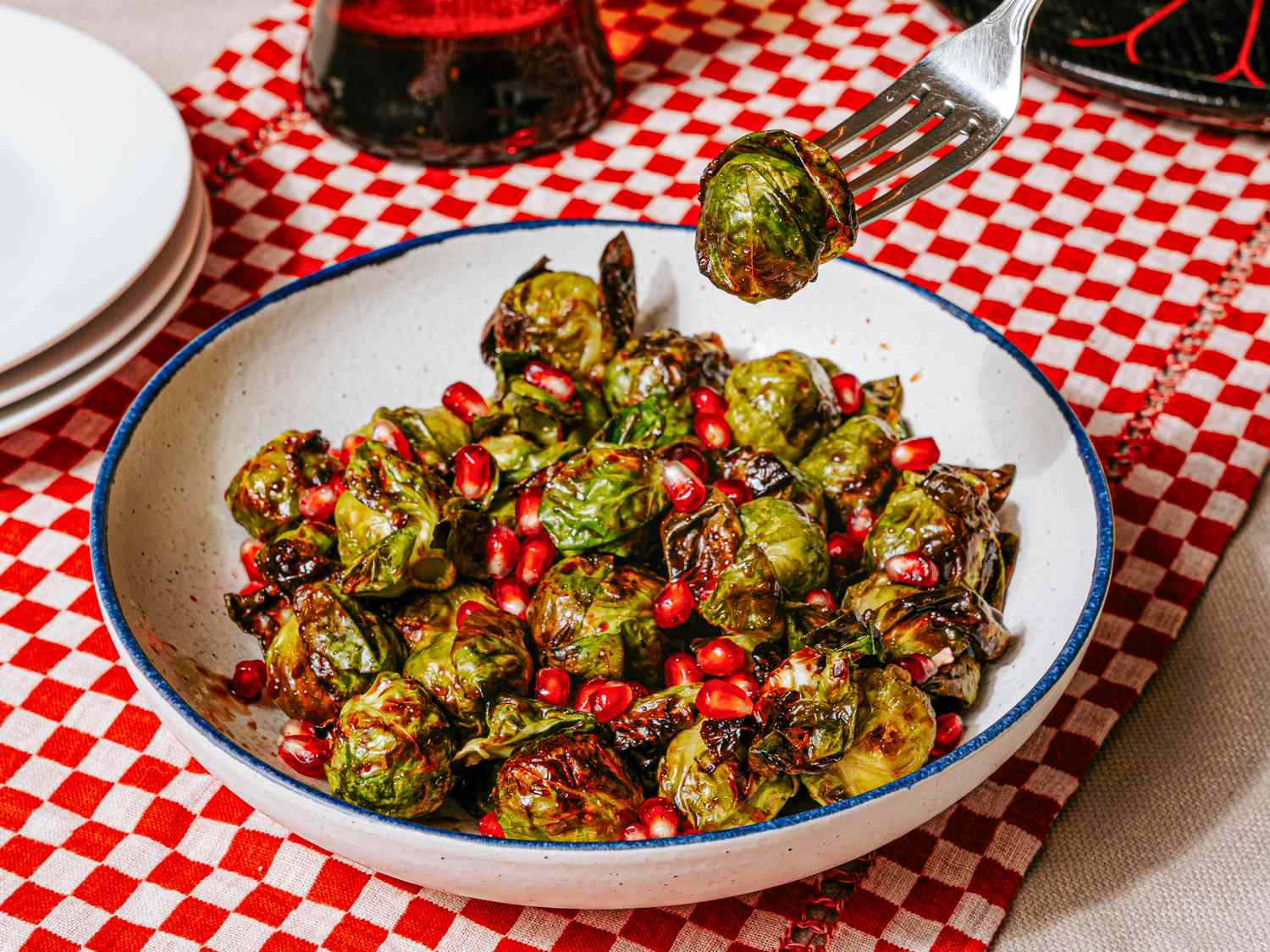 A bowl of airfried Brussels sprouts garnished with pomegranate seeds with a fork picking up a piece placed on a checkered tablecloth