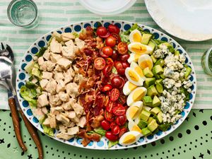 A Cobb salad served on a decorative platter, featuring rows of ingredients including chicken, bacon, cherry tomatoes, eggs, avocado, and blue cheese