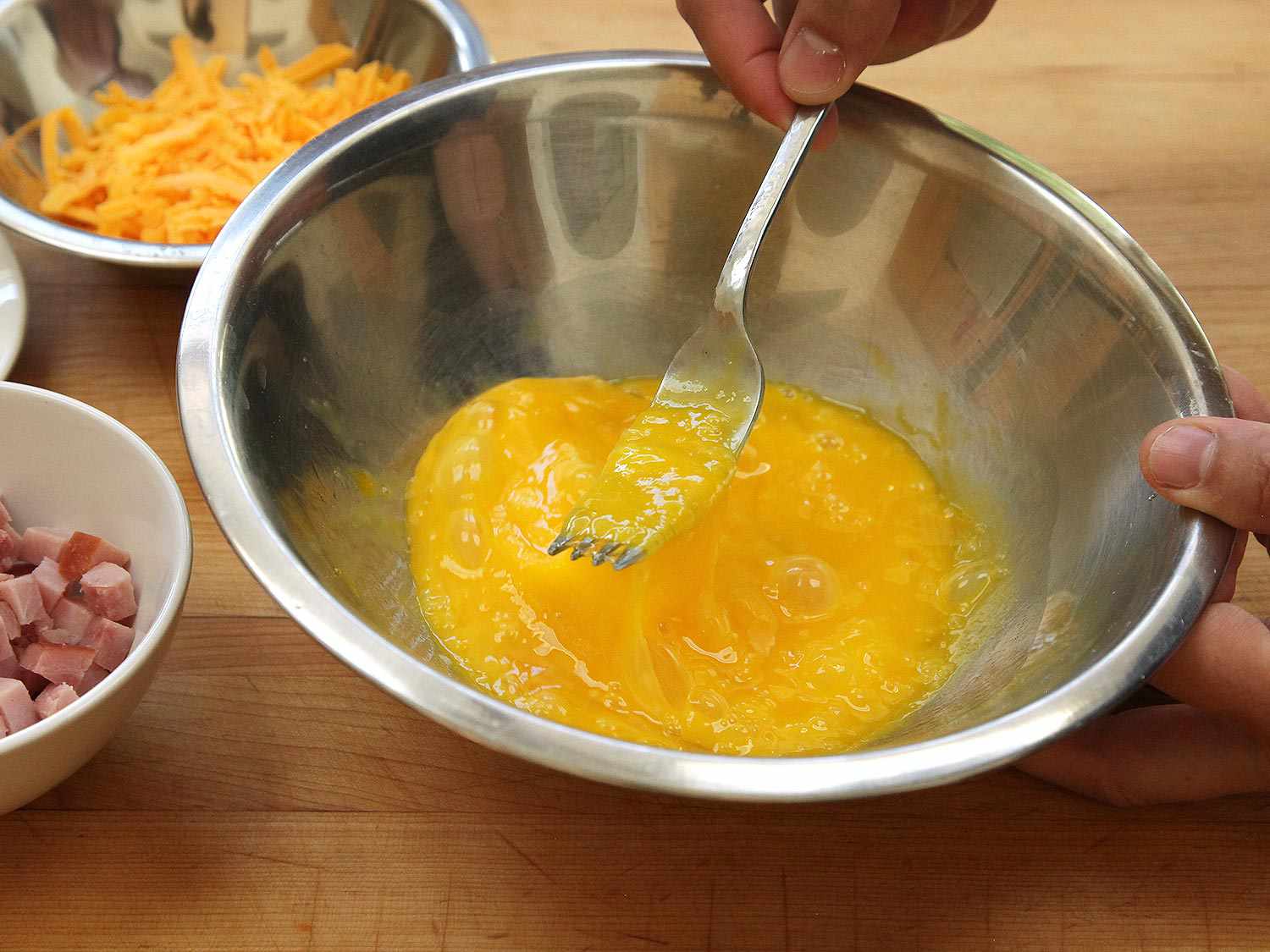 Eggs are beaten in a mixing bowl with a fork, bowl of grated cheddar cheese and another bowl of diced ham alongside.