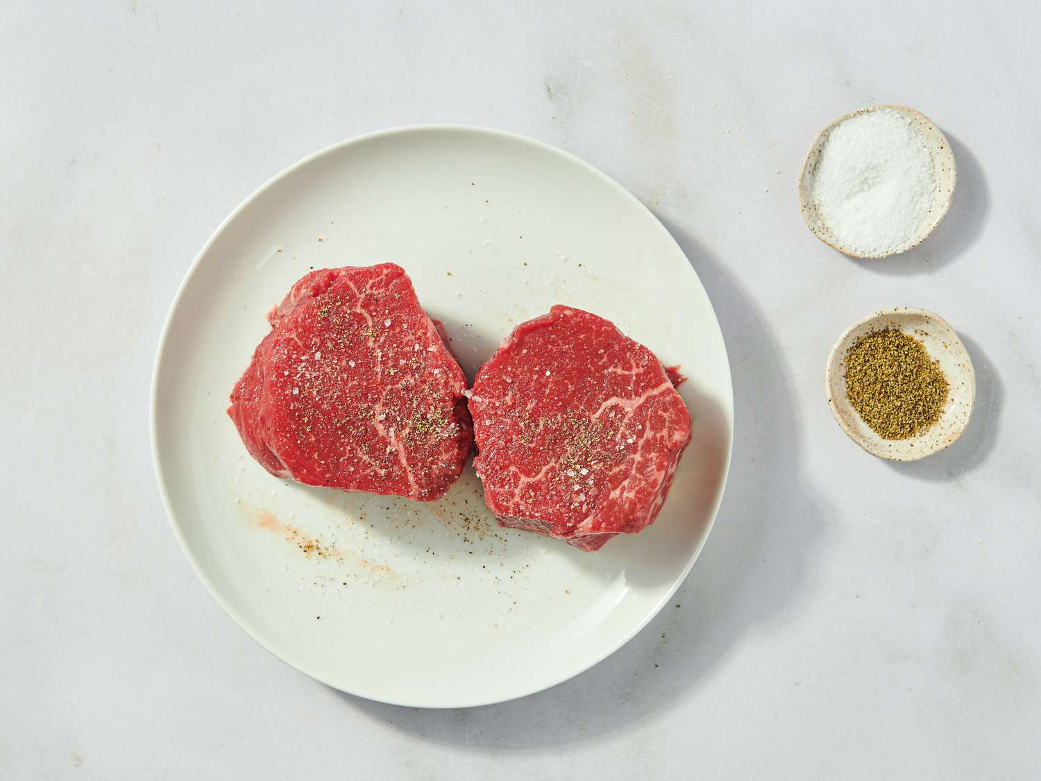 Two raw steaks on a white plate next to small bowls of salt and pepper