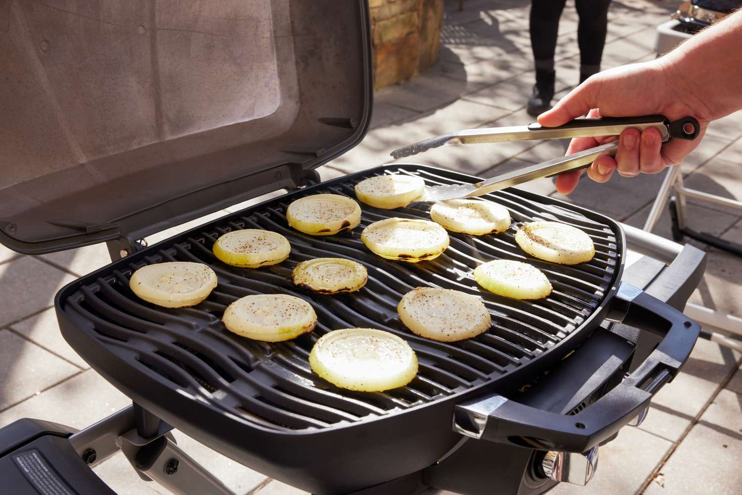 A person using tongs to flip onion rounds on a portable gas grill