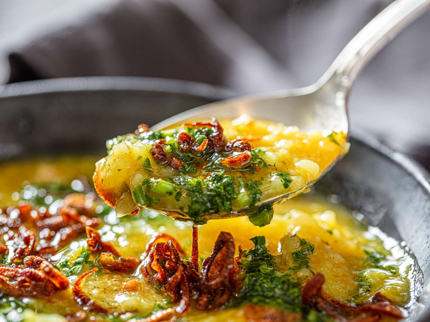 Closeup of a spoon lifting a portion of a dish with cooked rice garnished with herbs and fried shallots from a bowl