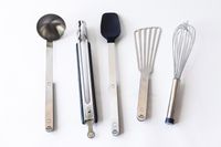 A set of five stainless steel kitchen utensils including a scoop tongs spatula slotted turner and whisk on a white surface
