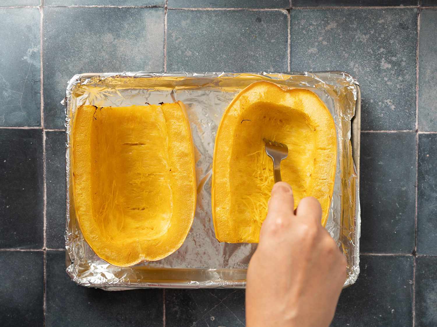 Roasted spaghetti squash halves on foil lined baking sheet being shredded with a fork