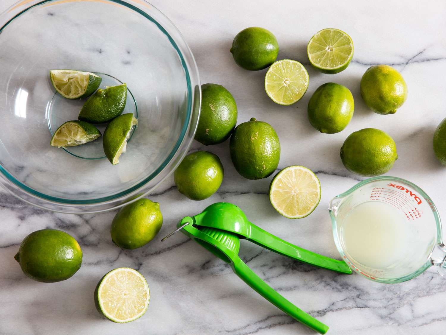 Overhead view of lime being juiced with a lever-style juice on a white marble work surface. A bowl of spent lime halves is reserved as the limes are juiced.