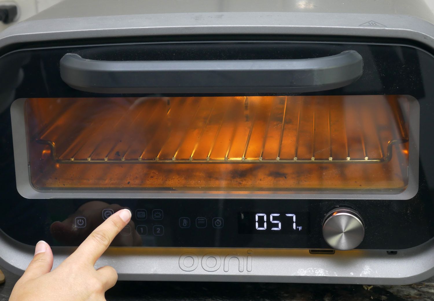 A hand pressing a button on a countertop oven with an interior light on and temperature displayed as 57