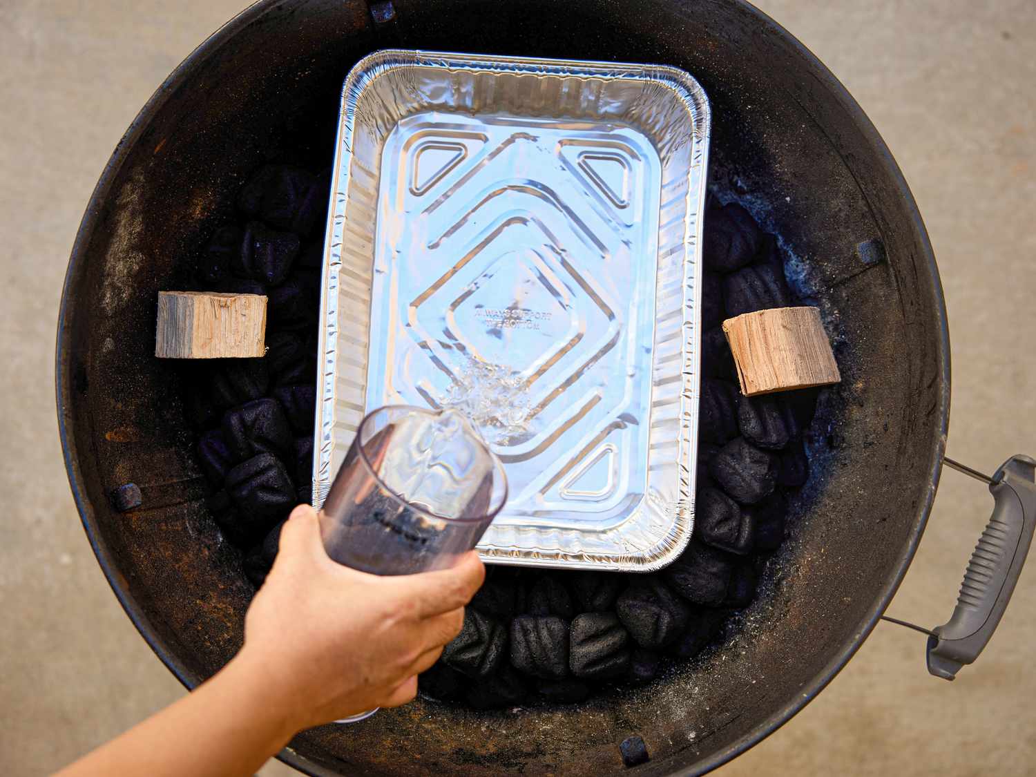 Pouring water into a foil pan inside a grill setup with charcoal and wood chunks on the sides
