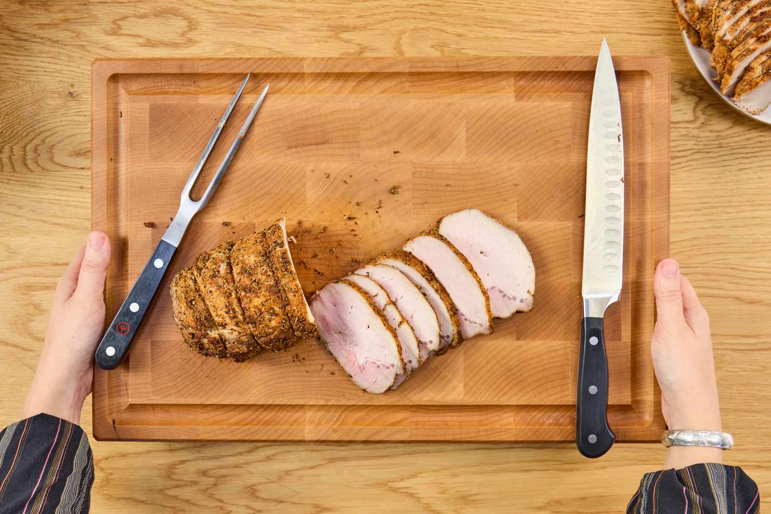 Hands holding a Boardsmith Maple End-Grain Cutting Board with sliced roast meat a carving fork and a knife