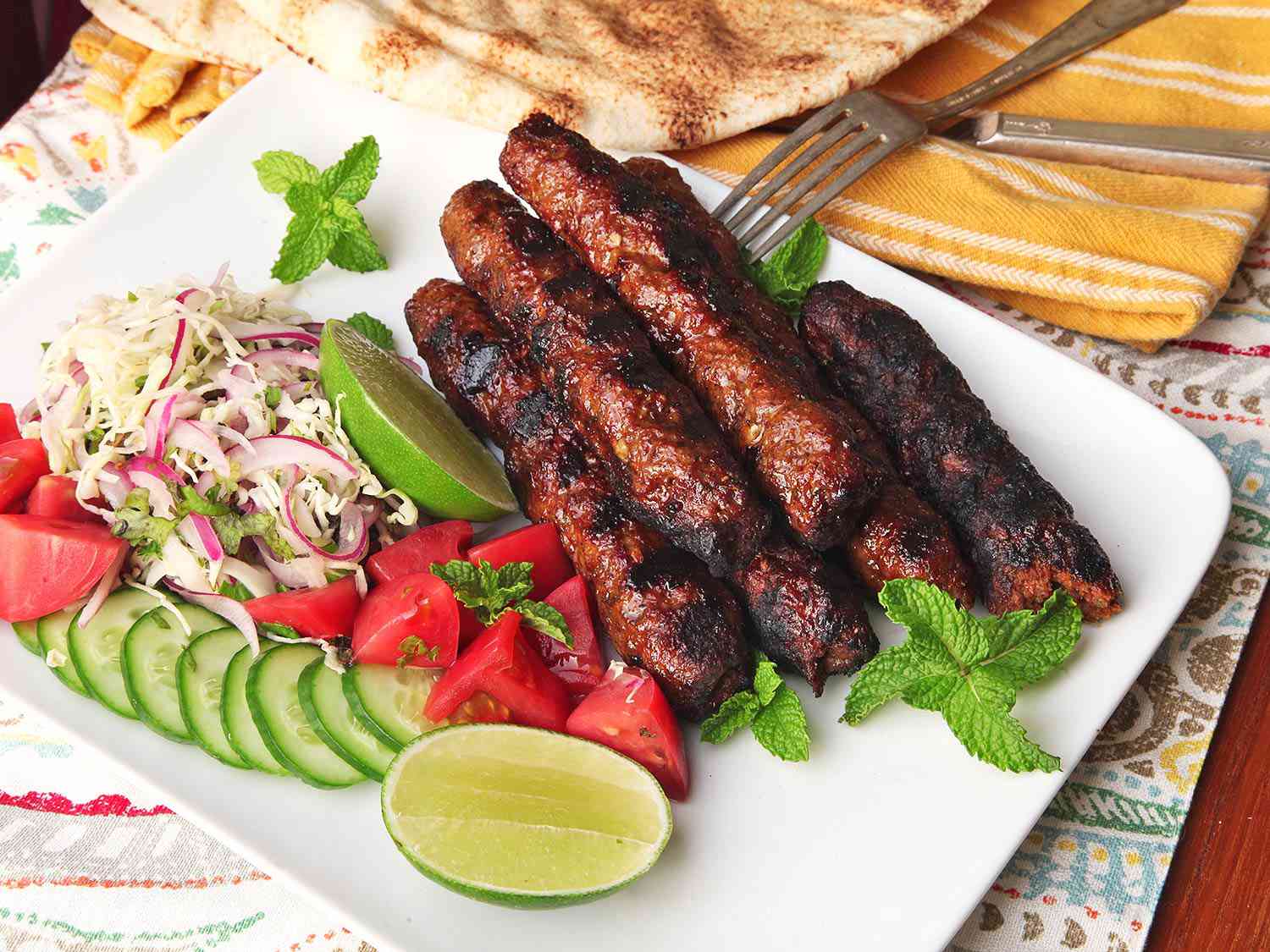 A platter of Pakistani Seekh kebabs with slaw, cucumbers, tomatoes, and lime wedges.