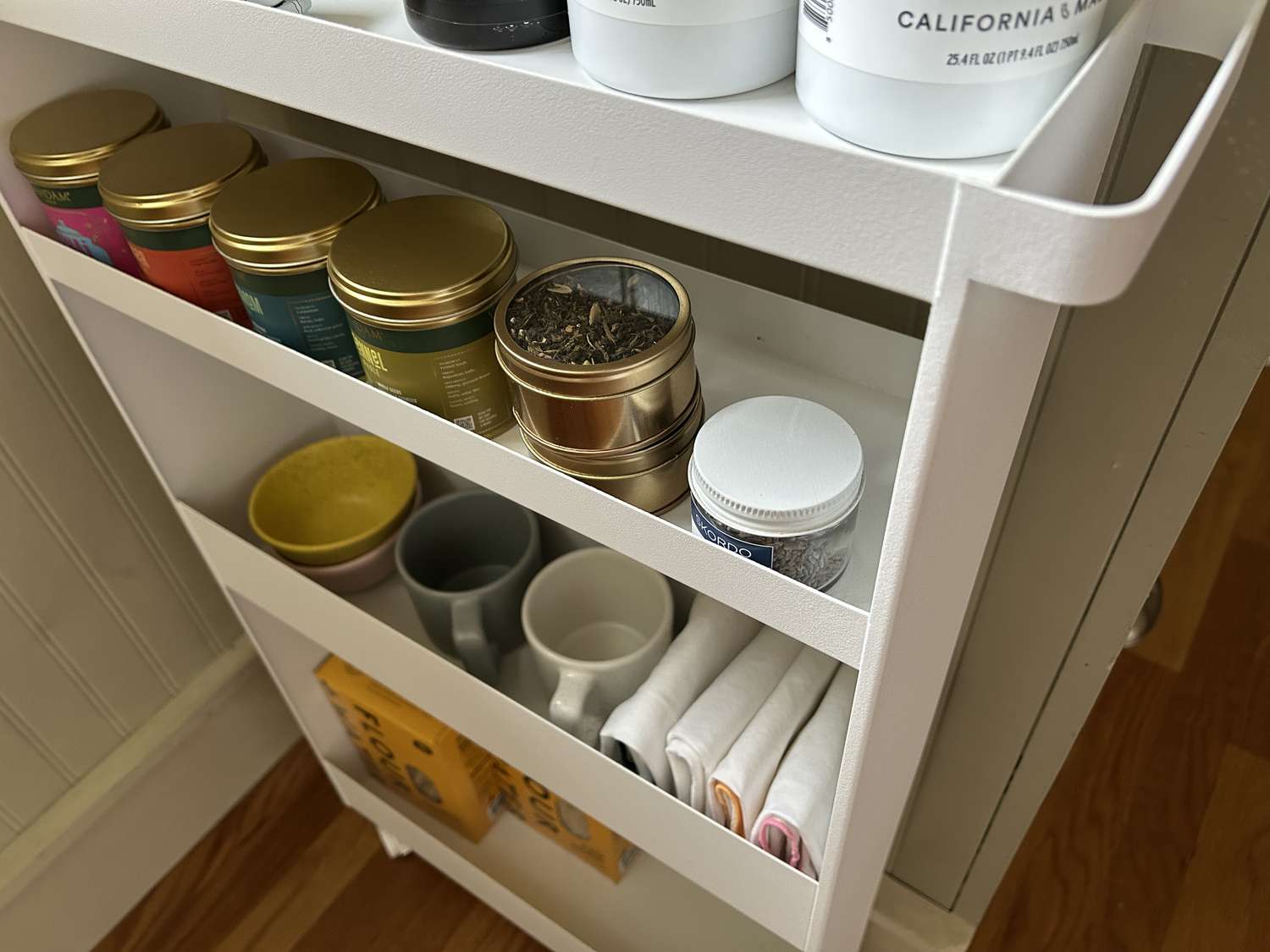 A white rolling kitchen storage rack with olive oil, spices, mugs, napkins, and boxes of pasta on its shelves