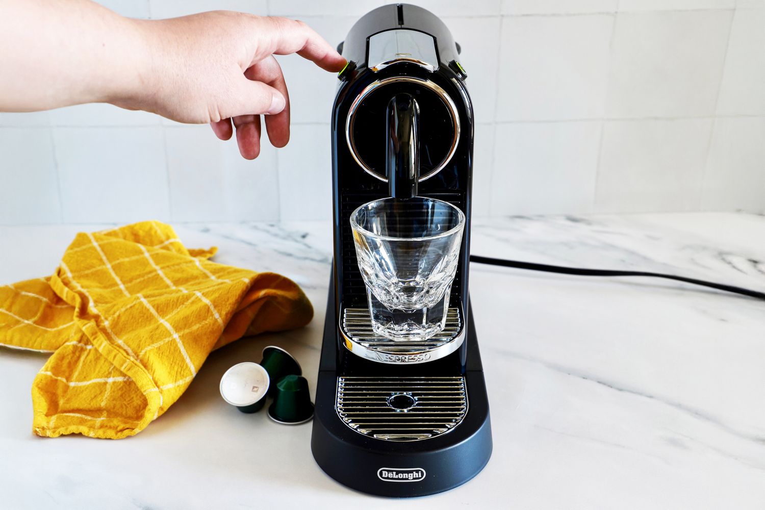 A hand operating a Nespresso espresso machine with an empty glass placed under the dispenser on a countertop