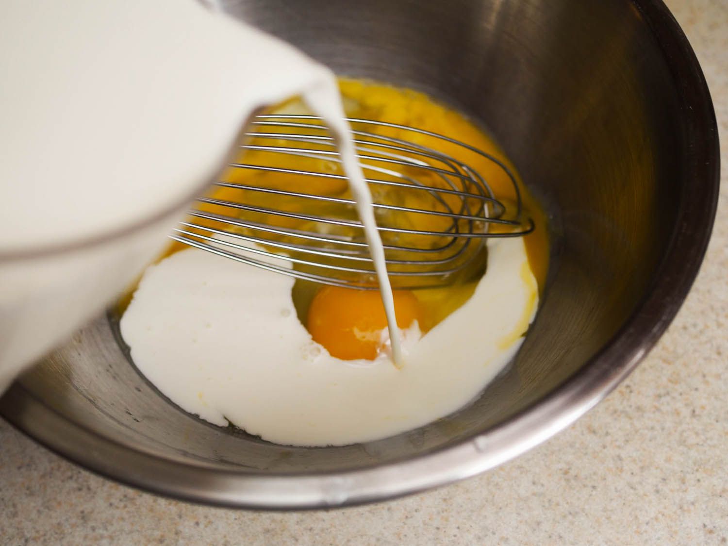 Cream is poured into a mixing bowl containing eggs (and a whisk).