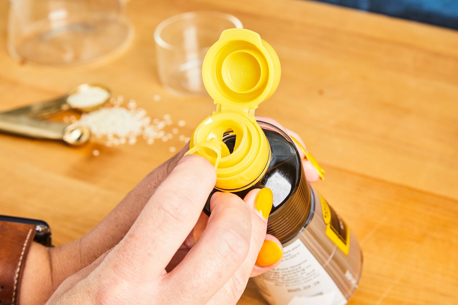 A hand holding and opening a bottle of liquid with a yellow cap, granules scattered on the countertop nearby