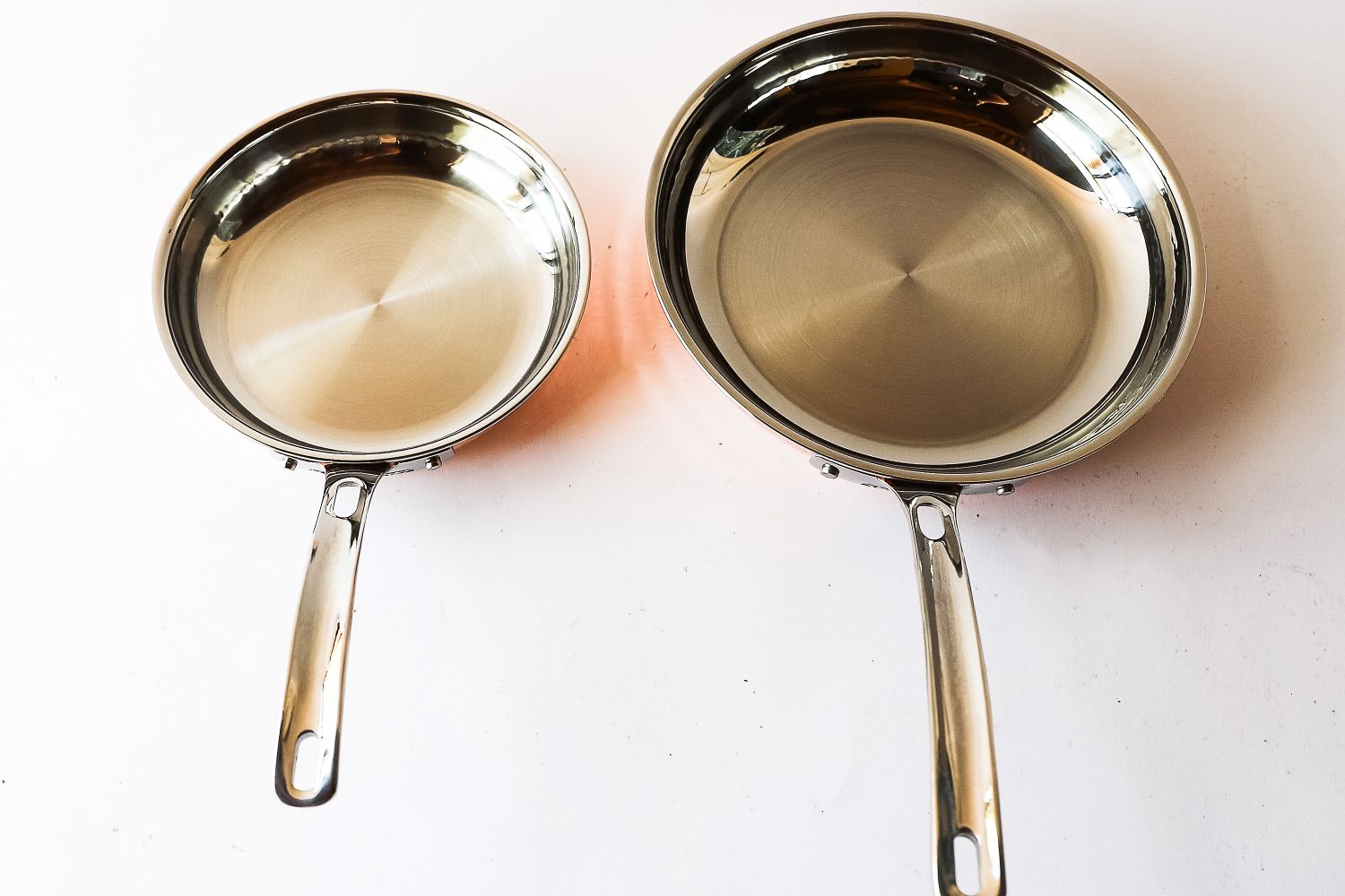 Two stainless steel frying pans one smaller and one larger placed side by side on a light surface showing their cooking surfaces and handles