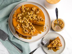 A deep orange sweet potato cheesecake drizzled with a flood of walnuts and caramel sits on a large plate with one slice missing; that slice is on a serving plate next to it.
