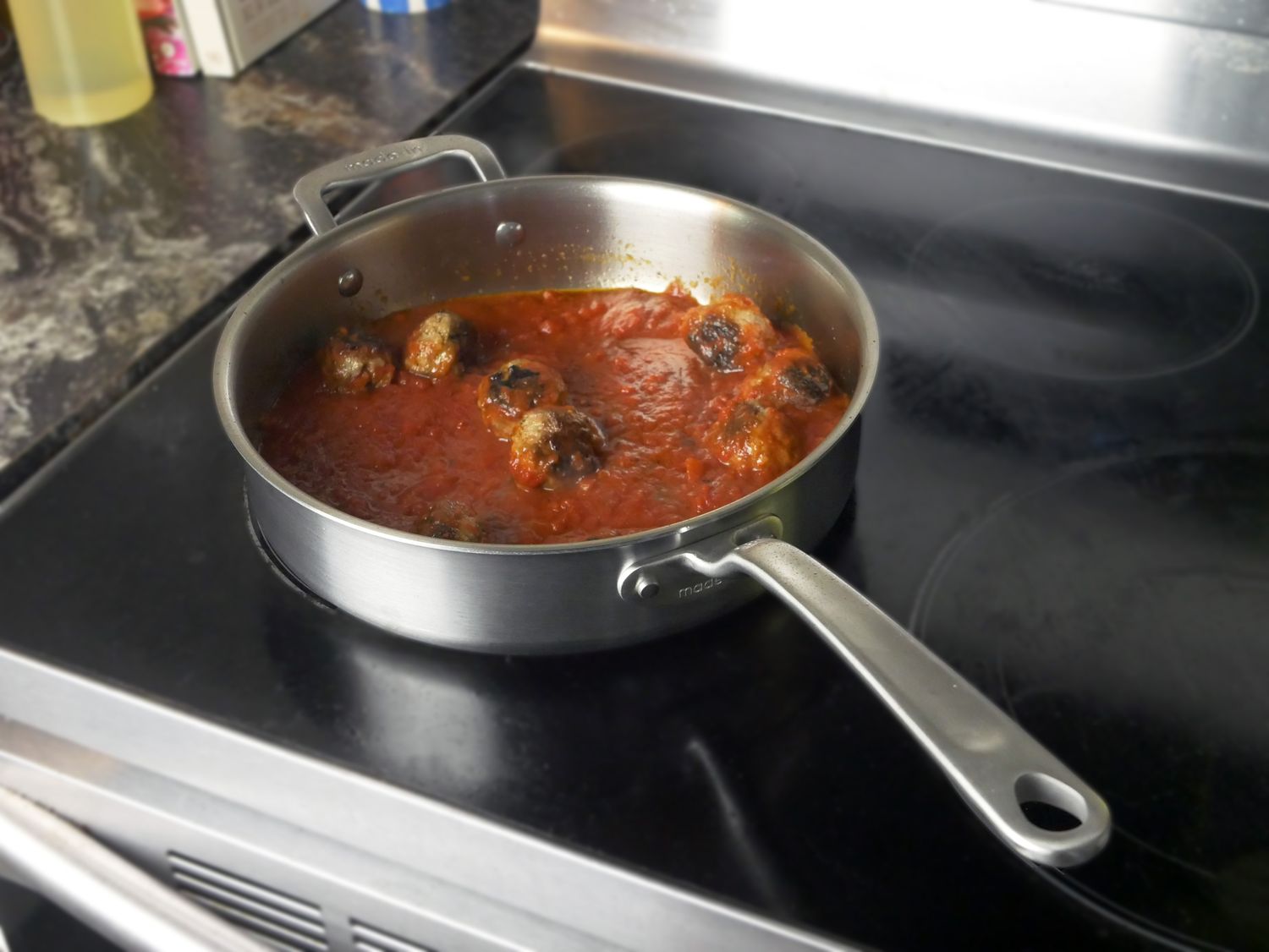a saute pan on a glass stovetop with meatballs and sauce inside.