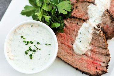A small ramekin of horseradish sauce on a plate of sliced roast beef with horseradish cream spooned over top. 